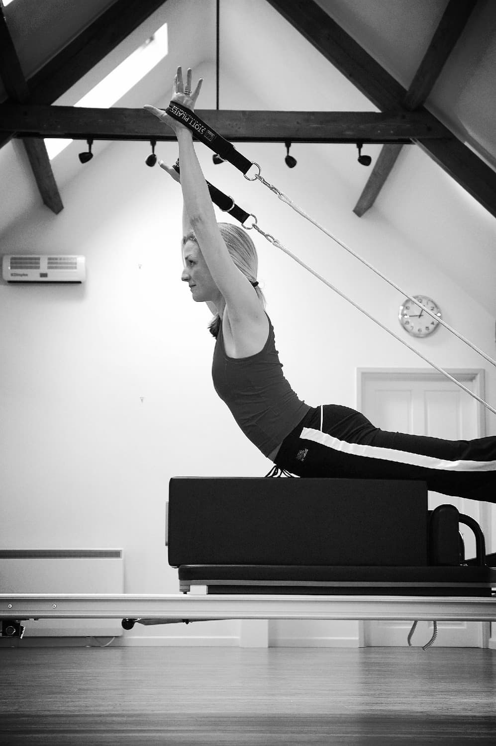 Kneeling on the reformer, both arms overhead on a ceiling-hung Stott Pilates strap, under the barn's oak beams